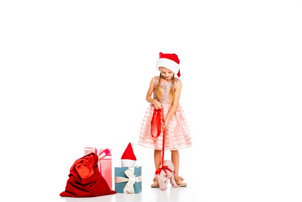 прелестный маленький ребенок в шляпе Санты с поводком поросенка и кучей рождественских подарков, изолированных на белом
 - Фото, изображение