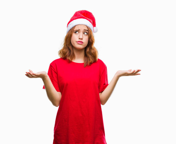 Молодая красивая женщина на изолированном фоне носит рождественскую шляпу невежественное и запутанное выражение лица с поднятыми руками и руками. Концепция сомнений
. - Фото, изображение