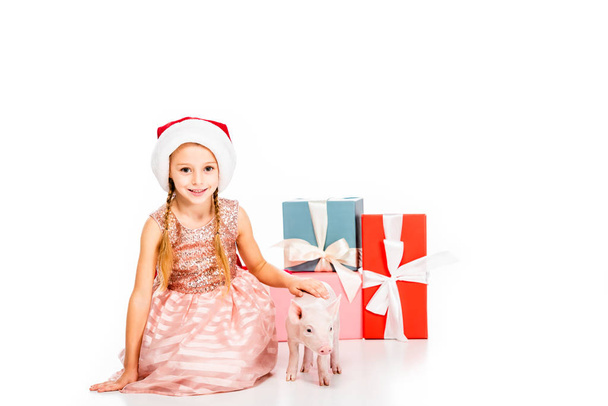 lindo niño feliz en santa sombrero acariciando cerdo y sonriendo a la cámara mientras está sentado cerca de regalos de Navidad aislado en blanco
 - Foto, Imagen