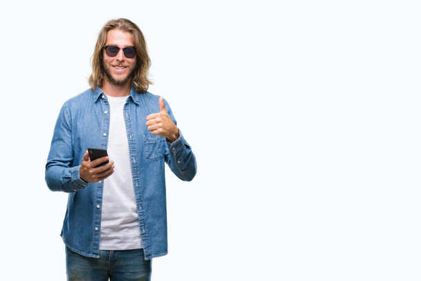 Young handsome man with long hair over isolated background sending message using smartphone happy with big smile doing ok sign, thumb up with fingers, excellent sign - Valokuva, kuva