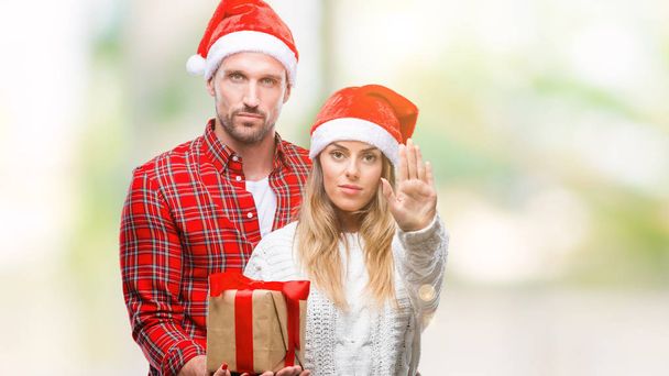 Молода пара закохана в різдвяний капелюх і тримає подарунок над ізольованим тлом з відкритою рукою робить знак зупинки з серйозним і впевненим виразом, жест захисту
 - Фото, зображення