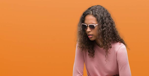 Beautiful young hispanic woman wearing sunglasses with hand on stomach because nausea, painful disease feeling unwell. Ache concept. - Foto, imagen