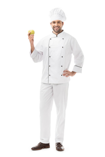 full length view of handsome young chef holding fresh apple and smiling at camera isolated on white - Valokuva, kuva