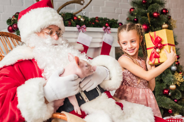 Santa spelen met varken en schattige lachende kind houden geschenkdoos  - Foto, afbeelding