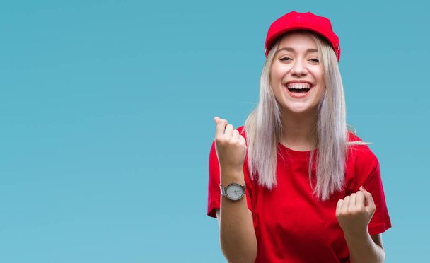 笑顔と成功のために叫んでで赤い帽子を身に着けている若いブロンドの女性は非常に幸せと興奮の腕を上げ、勝者のジェスチャを行う背景を分離しました。お祝いのコンセプト. - 写真・画像