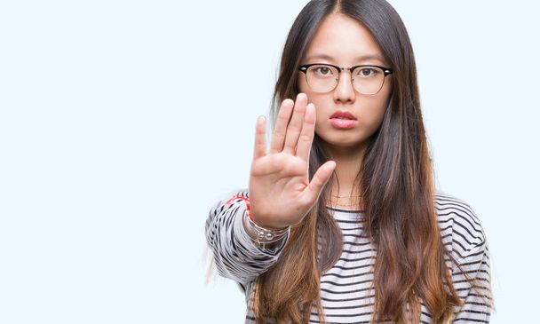 Молода азіатка в окулярах над ізольованим тлом припиняє співати з долонею. Попереджувальний вираз з негативним і серйозним жестом на обличчі
. - Фото, зображення