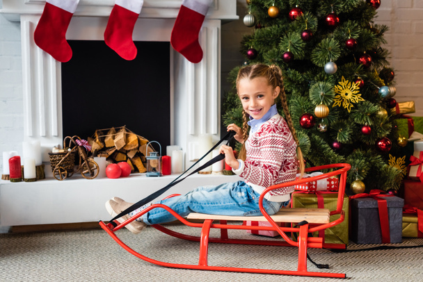 cute happy child sitting on sled and smiling at camera near fireplace - Foto, Bild