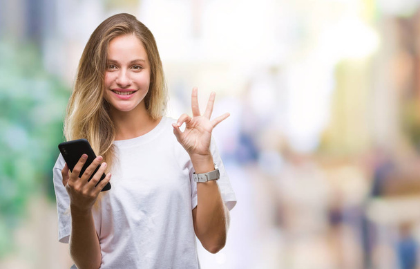 Young beautiful blonde woman using smartphone over isolated background doing ok sign with fingers, excellent symbol - Fotografie, Obrázek