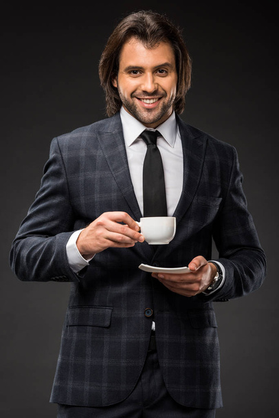 happy handsome businessman holding cup of coffee and smiling at camera isolated on black - Foto, Imagem