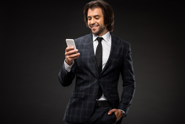 mosolygó fiatal üzletember állva a kéz a zsebben, és használ smartphone elszigetelt fekete - Fotó, kép