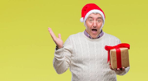 Handsome senior man wearing christmas hat and holding gift over isolated background very happy and excited, winner expression celebrating victory screaming with big smile and raised hands - Fotografie, Obrázek