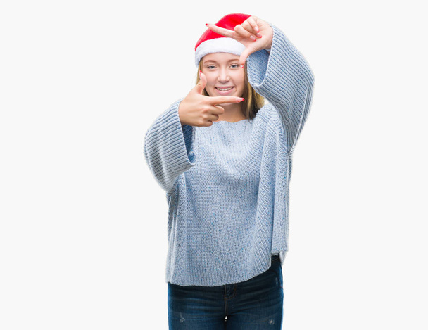 Joven hermosa mujer caucásica con sombrero de Navidad sobre fondo aislado sonriendo haciendo marco con las manos y los dedos con la cara feliz. Concepto de creatividad y fotografía
. - Foto, Imagen