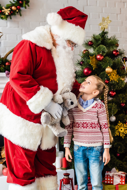 santa holding teddy bear and looking at cute smiling child standing near christmas tree - Photo, Image