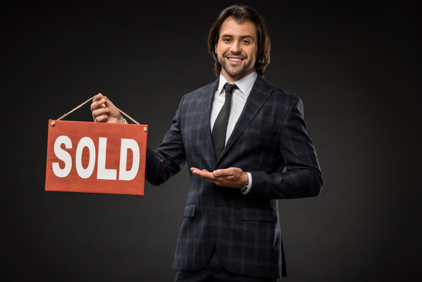 professional young businessman showing sold sign and smiling at camera isolated on black  - Fotó, kép