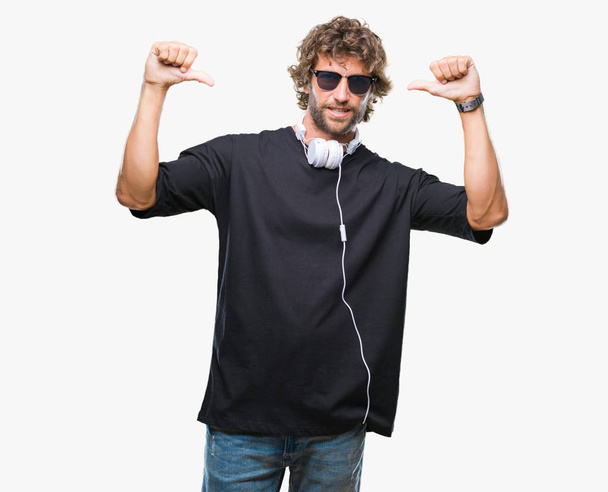 Hombre hispano guapo escuchando música usando auriculares sobre un fondo aislado que se ve confiado con sonrisa en la cara, señalándose con los dedos orgullosos y felices
. - Foto, Imagen