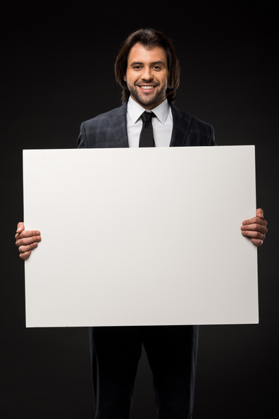 professional young businessman holding blank banner and smiling at camera isolated on black - Photo, Image