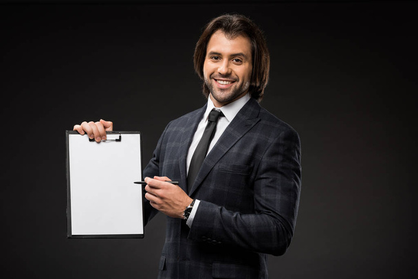 professional young businessman pointing at blank clipboard and smiling at camera isolated on black - Фото, изображение