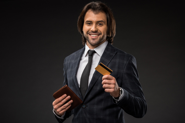 smiling young businessman holding wallet and credit card isolated on black  - 写真・画像