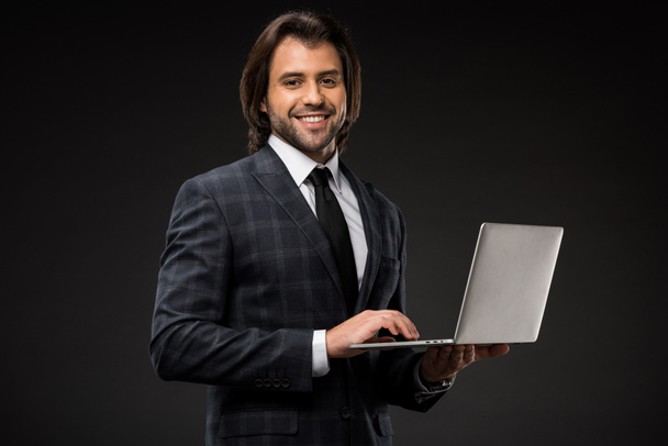 guapo joven hombre de negocios utilizando el ordenador portátil y sonriendo a la cámara aislada en negro
  - Foto, imagen