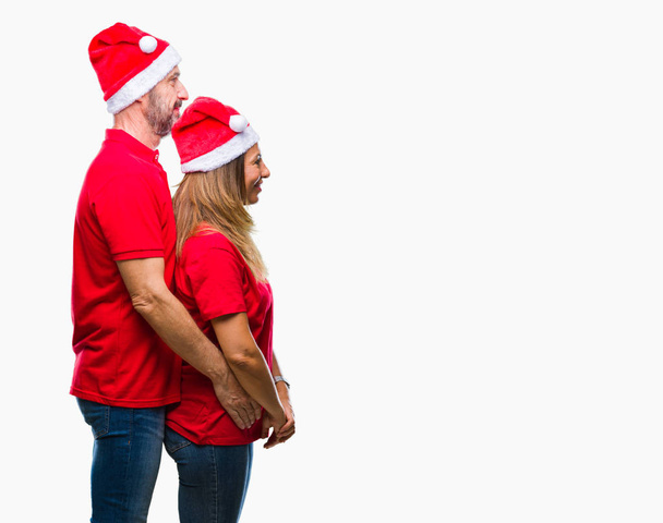 Μέση ηλικία φοράει καπέλο Χριστουγέννων πέρα από το απομονωμένο υπόβαθρο ψάχνει πλευρά, Ισπανόφωνος ζευγάρι χαλαρώσετε προφίλ πόζα με φυσικό πρόσωπο με αυτοπεποίθηση χαμόγελο. - Φωτογραφία, εικόνα