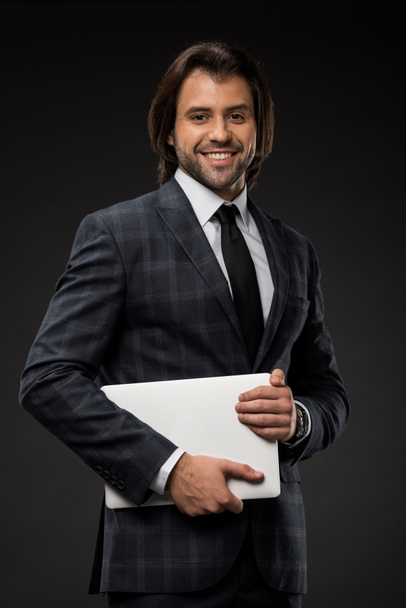handsome young businessman holding laptop and smiling at camera isolated on black  - Φωτογραφία, εικόνα