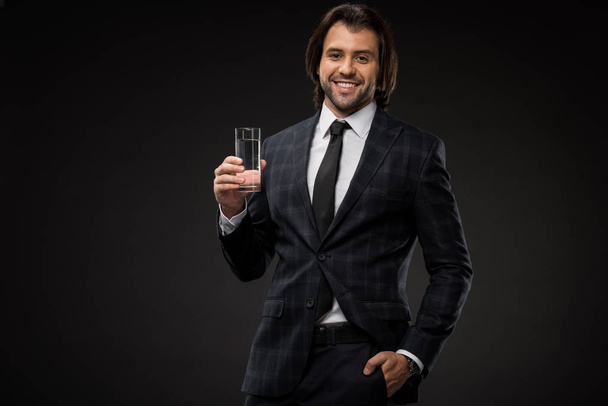 handsome young businessman holding glass of water and smiling at camera isolated on black - Φωτογραφία, εικόνα