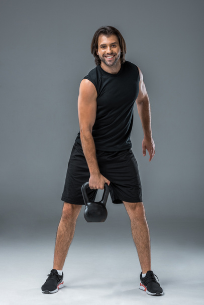 handsome sportsman holding kettlebell and smiling at camera on grey - Фото, изображение