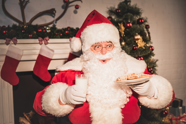 эмоциональный Санта держит тарелку с печеньем и стаканом молока
 - Фото, изображение