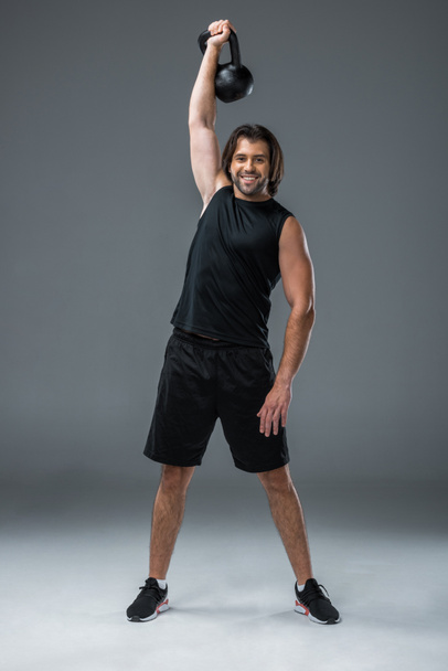 full length view of muscular young man training with kettlebell and smiling at camera on grey  - Photo, Image