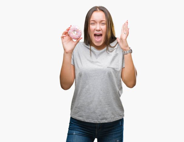 Молода біла жінка їсть солодкий пончик над ізольованим тлом дуже щасливий і схвильований, вираз переможця, який святкує перемогу, кричить з великою посмішкою і піднятими руками
 - Фото, зображення