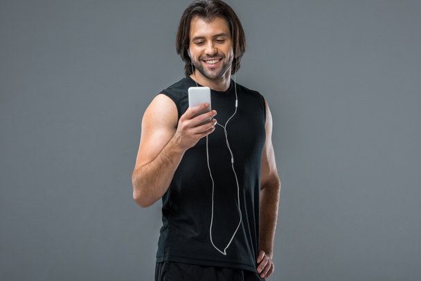 Lächelnder junger Sportler mit Smartphone und Musik in Kopfhörern isoliert auf grau - Foto, Bild