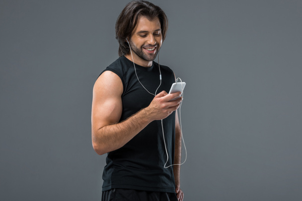 hombre musculoso sonriente en auriculares usando teléfono inteligente aislado en gris
 - Foto, imagen