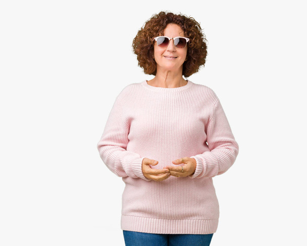 Hermosa mujer de mediana edad ager vistiendo suéter rosa y gafas de sol sobre fondo aislado Las manos juntas y los dedos cruzados sonriendo relajado y alegre. Éxito y optimismo
 - Foto, Imagen