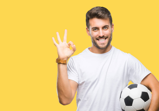 Mladý pohledný muž drží fotbal fotbalový míč nad izolované pozadí dělá ok cedulka s prsty, vynikající symbol - Fotografie, Obrázek