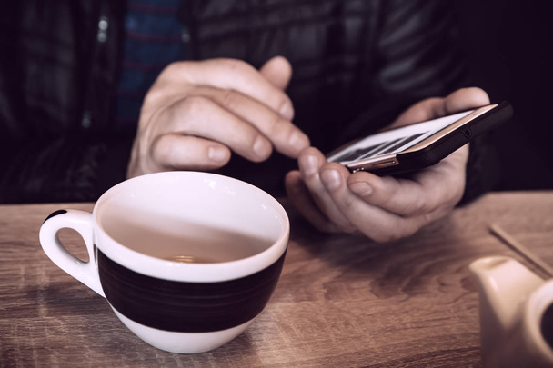 A man is resting in a cafe, drinking tea, reading and writing messages on a smartphone. Work in the mobile office. Close-up soft focus image with shallow depth of field. Old style vintage color - Photo, Image