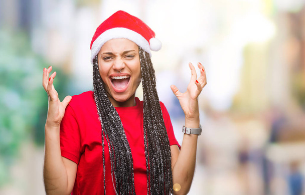 Joven chica afroamericana trenzada con sombrero de navidad sobre un fondo aislado celebrando loco y loco por el éxito con los brazos levantados y los ojos cerrados gritando emocionados. Concepto ganador
 - Foto, Imagen