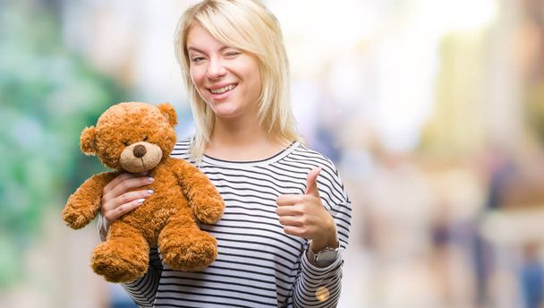 Молодая красивая блондинка с плюшем плюшевого медведя на изолированном фоне счастлива с большой улыбкой делает хорошо знак, палец вверх пальцами, отличный знак
 - Фото, изображение