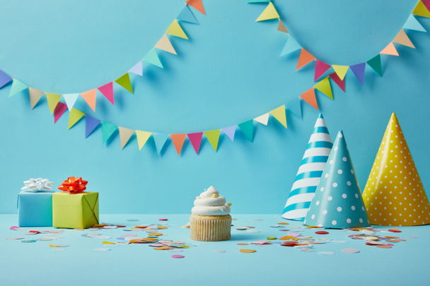  Sabroso cupcake, sombreros de fiesta, confeti y regalos sobre fondo azul con colorido bunting
 - Foto, Imagen
