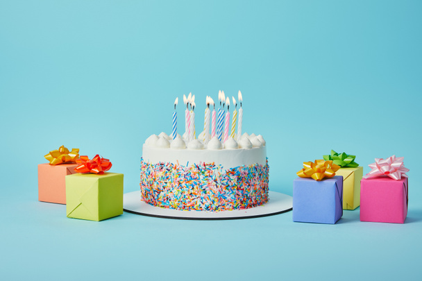 Heerlijke cake met kaarsen, kleurrijke geschenken en confetti op blauwe achtergrond - Foto, afbeelding