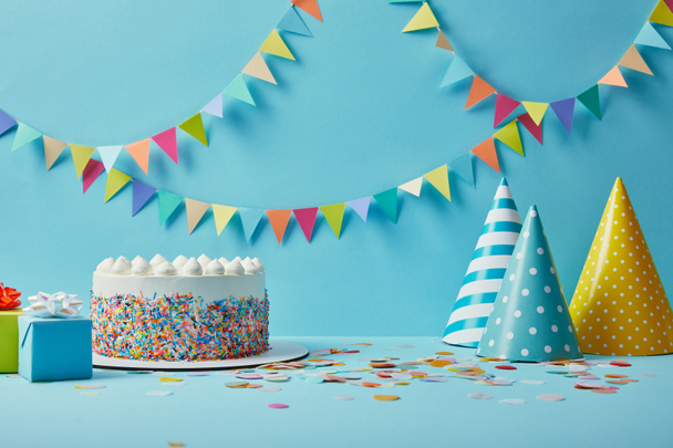 Tasty cake with sugar sprinkles,party hats and gifts on blue background with colorful bunting - Photo, Image