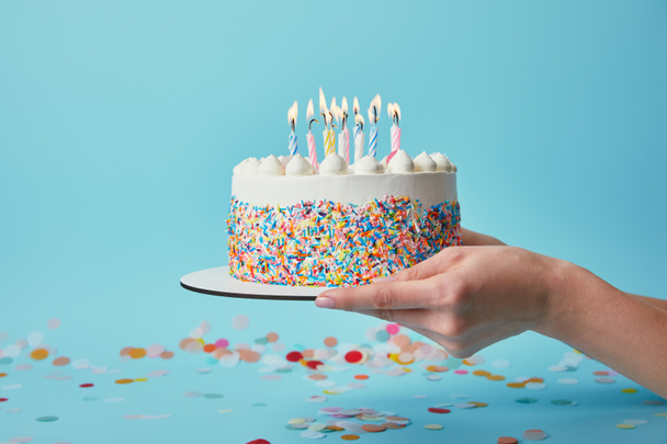 Vista parcial de la mujer sosteniendo pastel de cumpleaños con velas sobre fondo azul con confeti
 - Foto, imagen