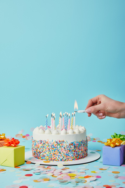 Partial view of woman lighting birthday cake on blue background with gifts and confetti - Fotoğraf, Görsel