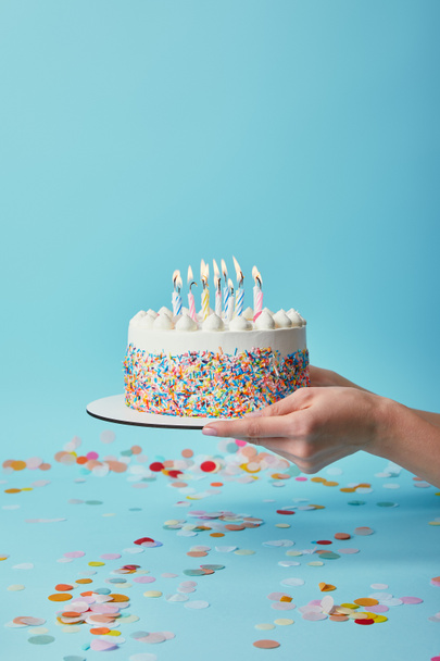 Vista recortada de la mujer sosteniendo delicioso pastel de cumpleaños con velas sobre fondo azul con confeti
 - Foto, Imagen