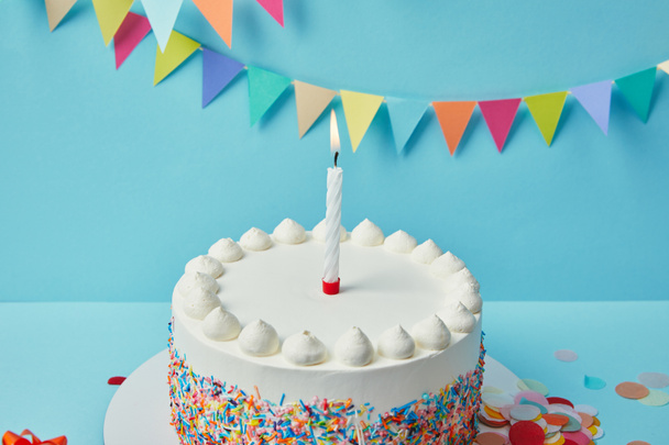 Candle on birthday cake with sugar sprinkles on blue background with bunting - Photo, Image