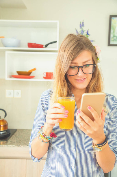 Χαριτωμένο κορίτσι πίνοντας φρέσκο χυμό πορτοκάλι και χρησιμοποιώντας το κινητό στην κουζίνα. - Φωτογραφία, εικόνα