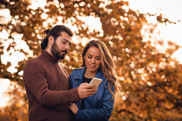 Young couple using cellphone in autumn colored park. - Foto, imagen