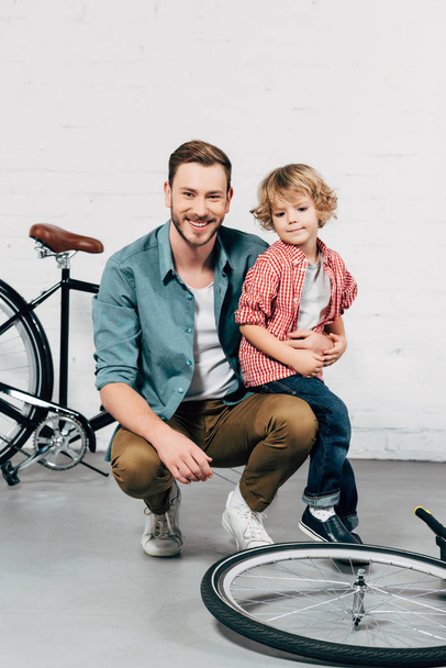 cheerful handsome man sitting with little son and looking at camera in bicycle workshop - Фото, изображение
