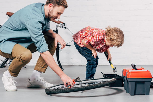 Seitenansicht eines männlichen Reparateurs, der ein Rad mit einer Zange fixiert, während sein kleiner Sohn mit Werkzeugkiste in der Werkstatt steht - Foto, Bild