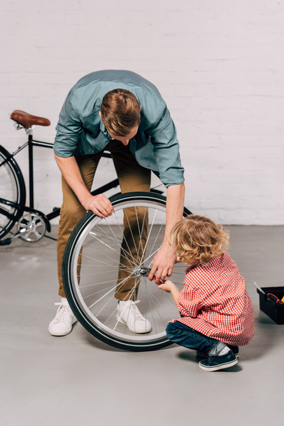 man helping little son repairing bicycle wheel with pliers at workshop - Фото, изображение