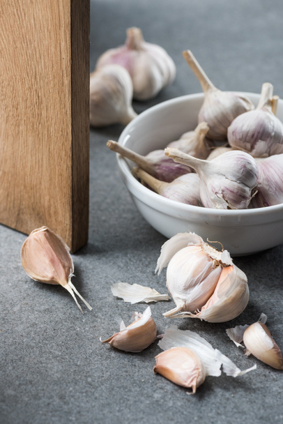 Garlic in ceramic bowl, several peeled cloves and wooden chopping board on grey background  - Фото, изображение